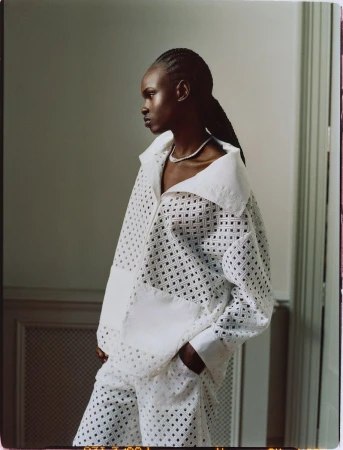 Portrait of a woman in an airy, white patterned jacket and matching pants, set against a simple light-toned indoor background.