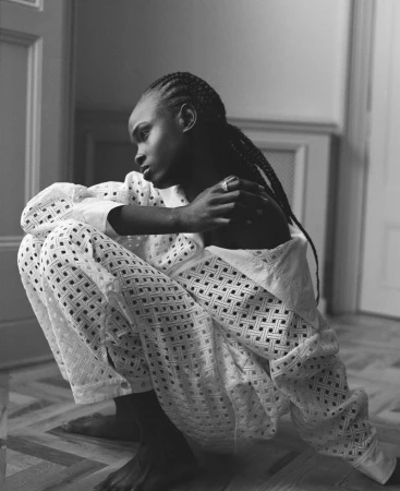 Portrait of an adult woman with dark skin and braids, posing indoors wearing a white, patterned outfit while crouching on a parquet floor.