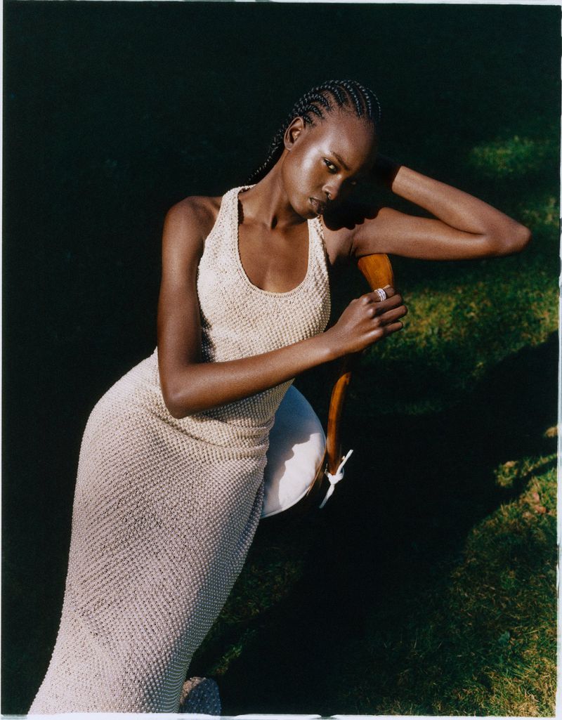 Model Bakhita Lual in a cream knit dress leaning against a wooden chair outdoors for Harper's Bazaar UK