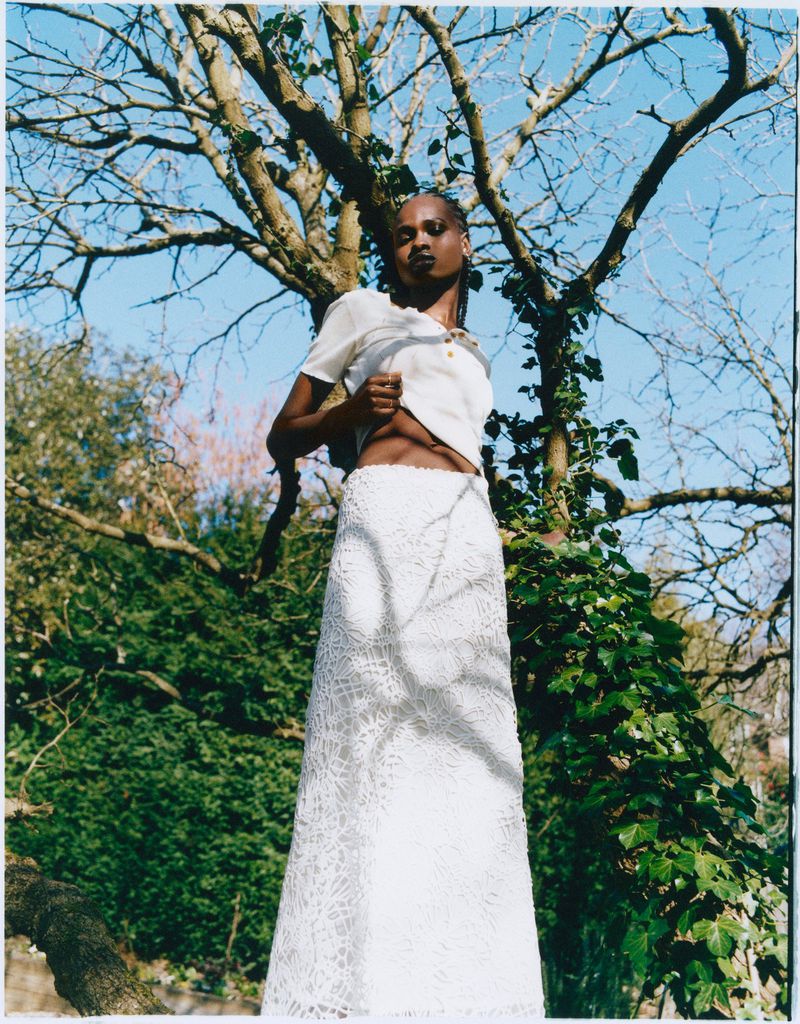 A black female model stands in a white lace skirt and top, posing outdoors among the trees and green foliage on a bright day.