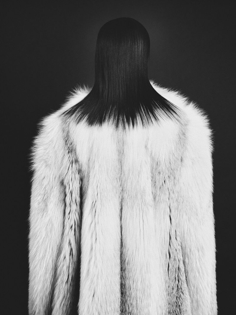Monochromatic fashion shot of a model wearing a long, white fur coat and slicked-back dark hair in a studio setting against a dark background.