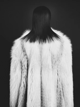 Monochromatic fashion shot of a model wearing a long, white fur coat and slicked-back dark hair in a studio setting against a dark background.