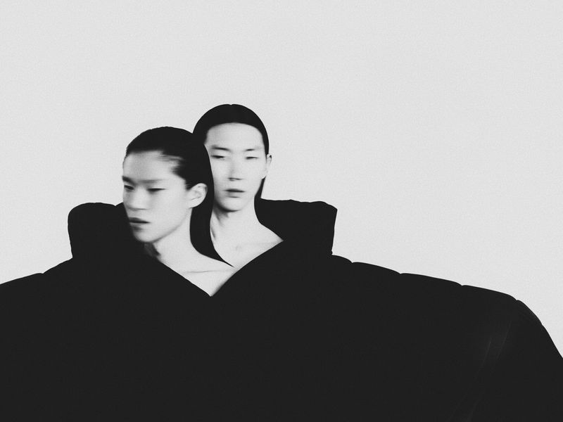 Monochrome portrait of two young Asian adults wearing black, draped fabric against a solid white backdrop in a studio setting.
