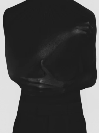 Close-up studio shot of an adult hugging themself; the composition highlights their bare back and arm skin, set against a plain background.
