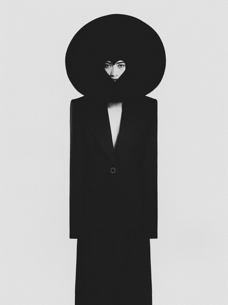 Fashion photography featuring a woman in a striking black outfit with a large, round hat against a minimalist light grey background.
