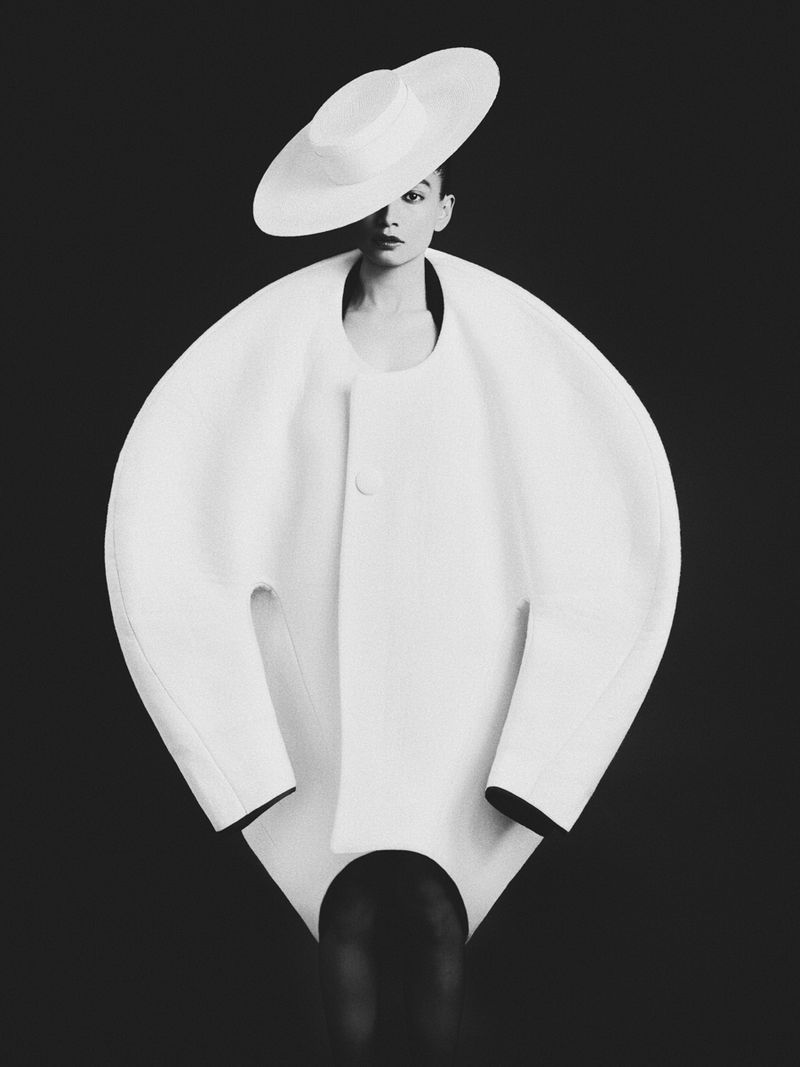 A woman poses in a sculptural, oversized white coat and matching hat against a black backdrop in this studio fashion portrait.