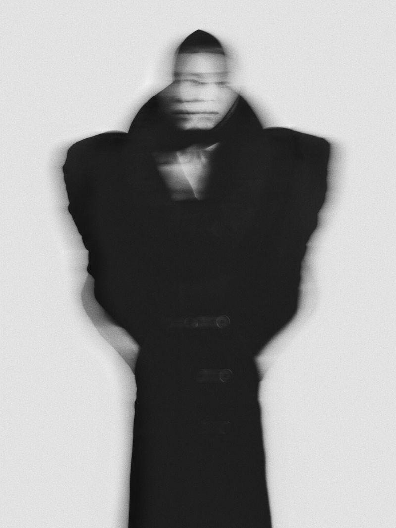 Monochromatic long-exposure portrait of a person in a dark coat against a white backdrop, with intentional motion blur creating a ghosting effect.