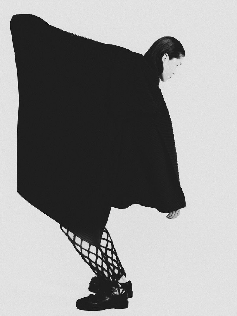 Monochrome fashion portrait of an androgynous person wearing a large, dark, shapeless dress with patterned tights and platform shoes against a white backdrop.