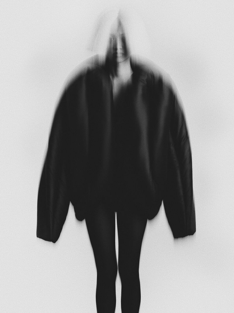Monochrome studio shot of a woman with a blurred effect, wearing a jacket, standing in front of a white wall with minimalist styling.