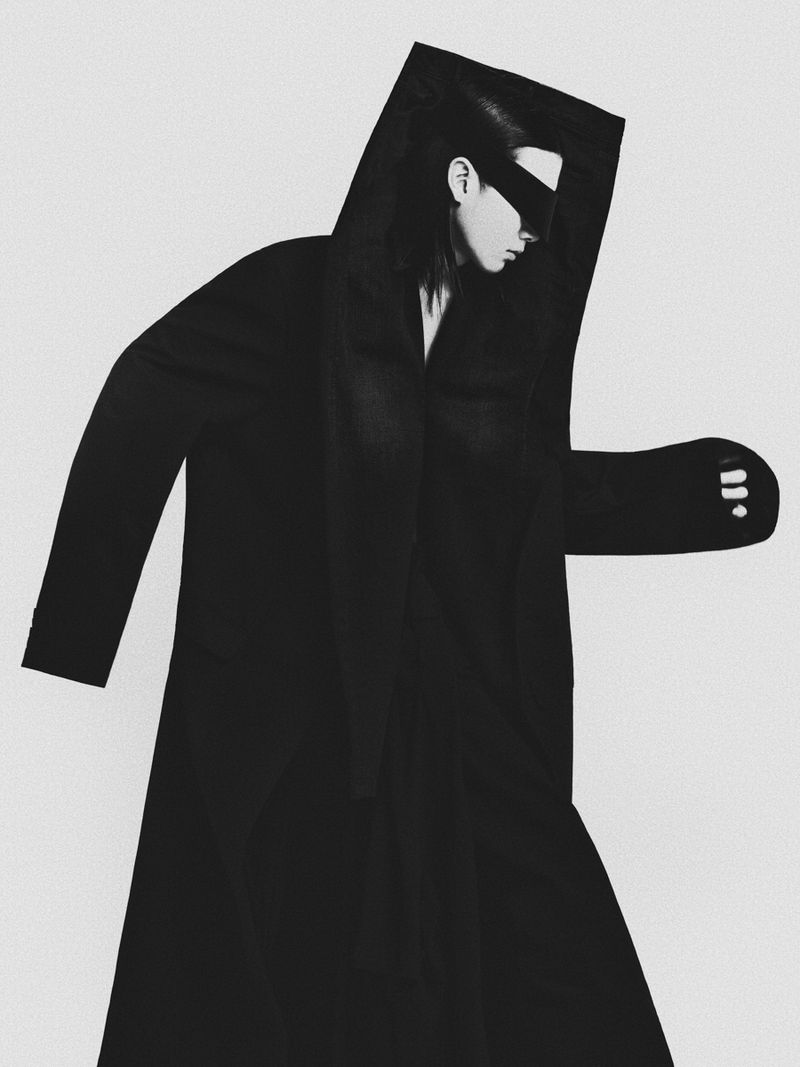Monochrome studio shot of a person wearing a black hooded coat and visor, standing against a plain white backdrop in a stylized pose.