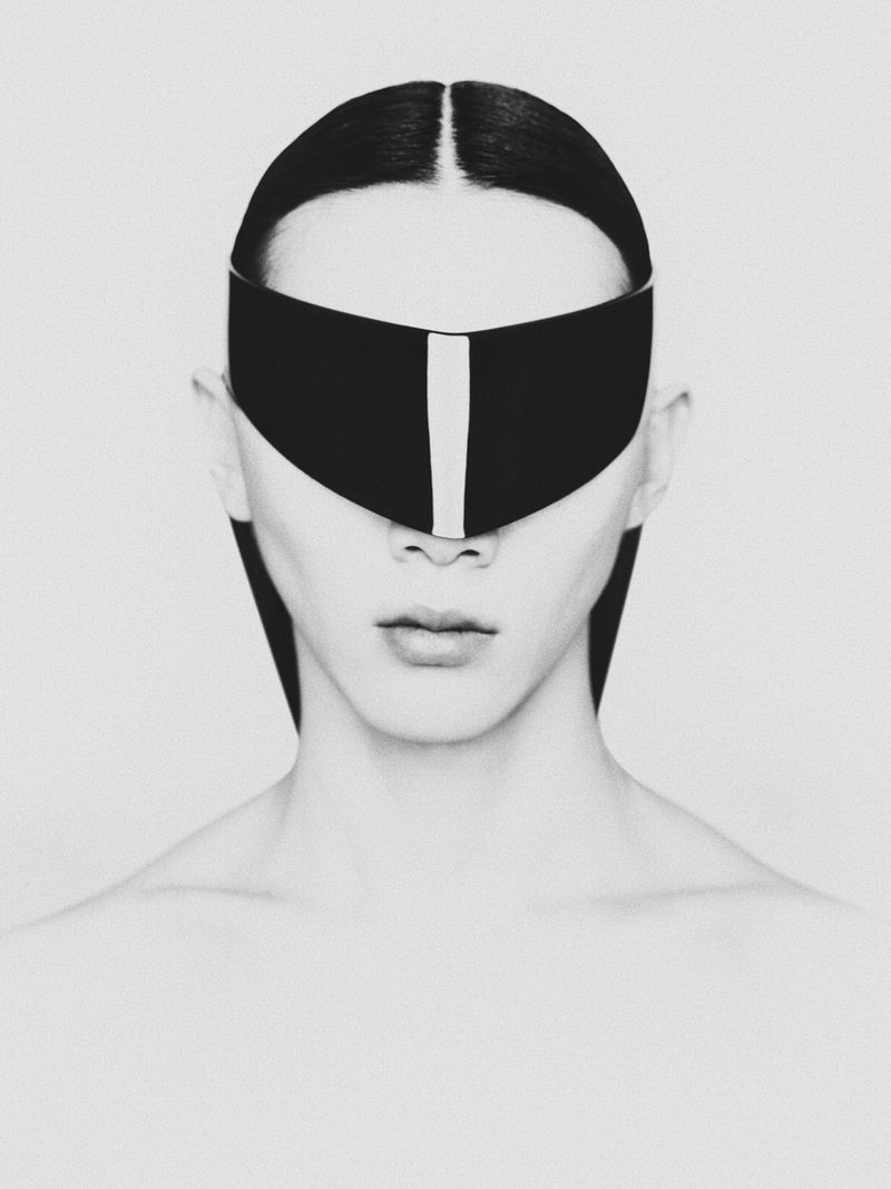 Striking black and white portrait of a person with dark hair and eyes obscured by a solid black visor, minimalist style, stark lighting.