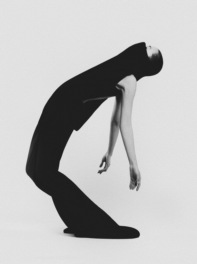 Monochromatic portrait of a woman in a dark hood and draped garment, gracefully arching her back in a minimalist studio setting.