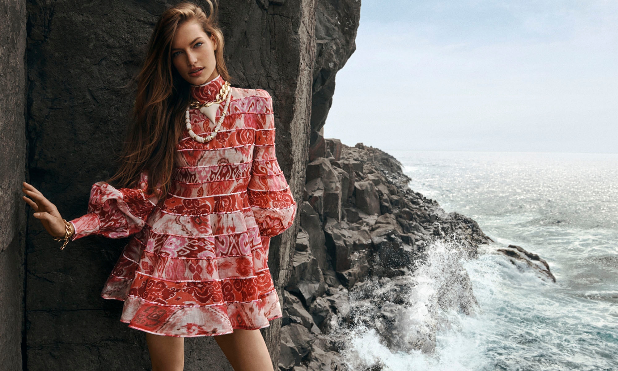 A woman in a red floral dress poses against a dark rock formation by the ocean with crashing waves in an outdoor fashion shoot.
