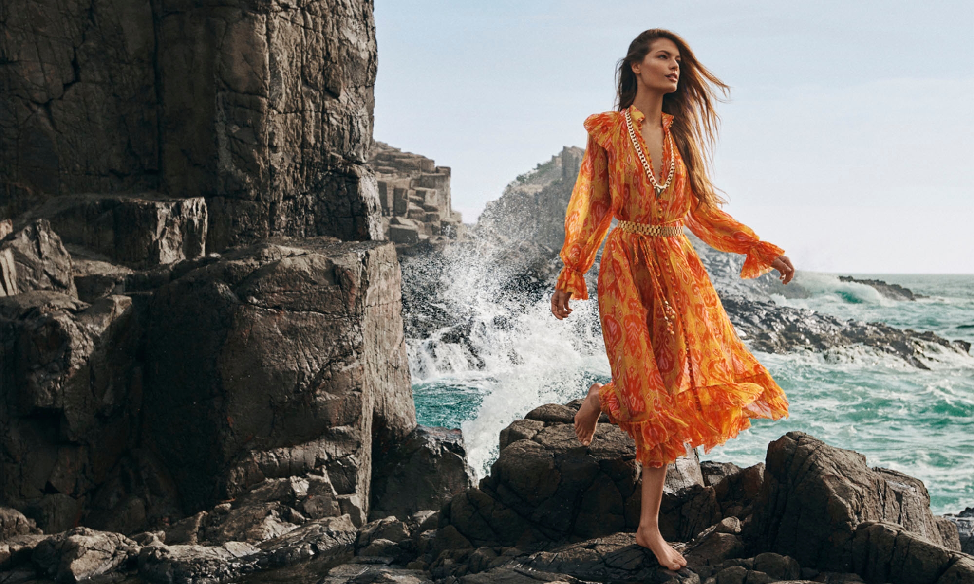 A woman in an orange floral dress poses barefoot on rocks by the ocean, featuring dramatic coastal cliffs and crashing waves.