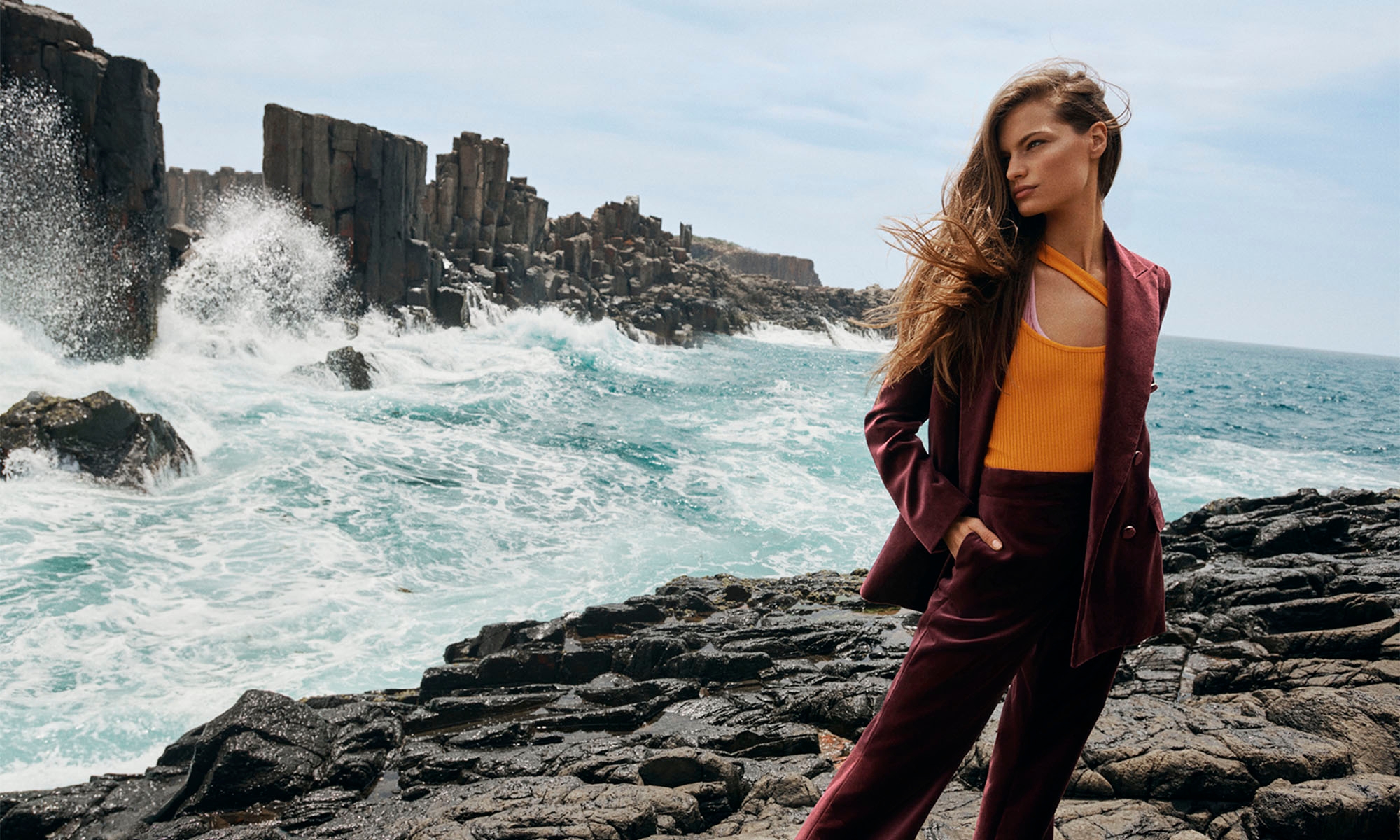 A woman in a suit stands on a rocky shoreline with dramatic rock formations and ocean waves in the background on a sunny day.