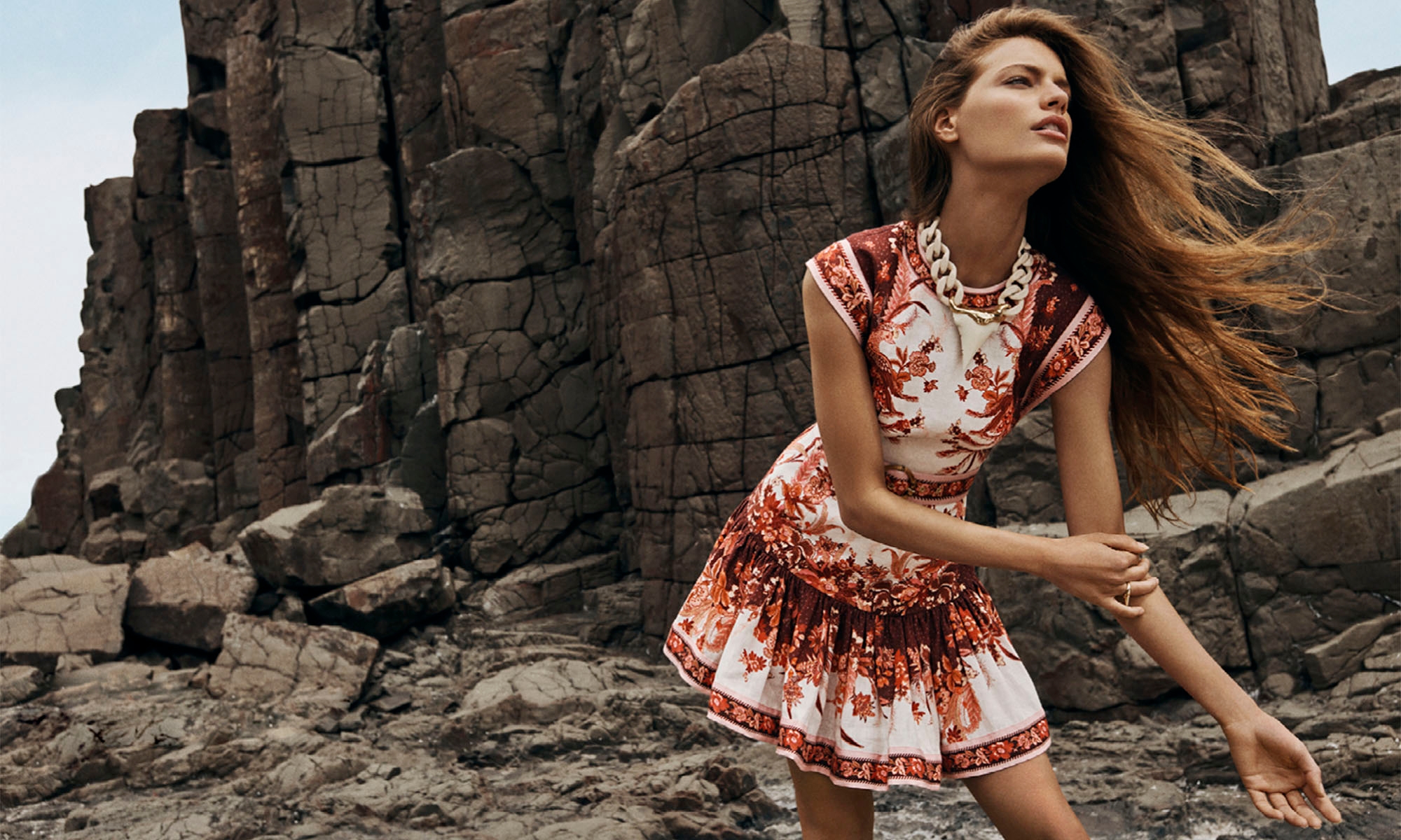 A woman in a patterned dress and necklace poses on a rocky beach with a unique rock formation in the background under natural light.