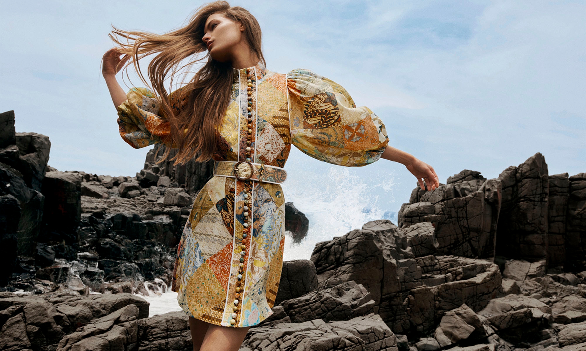 A fashionable adult woman in a patterned dress poses among rocky formations with ocean waves, under a bright sky, creating a vivid outdoor scene.