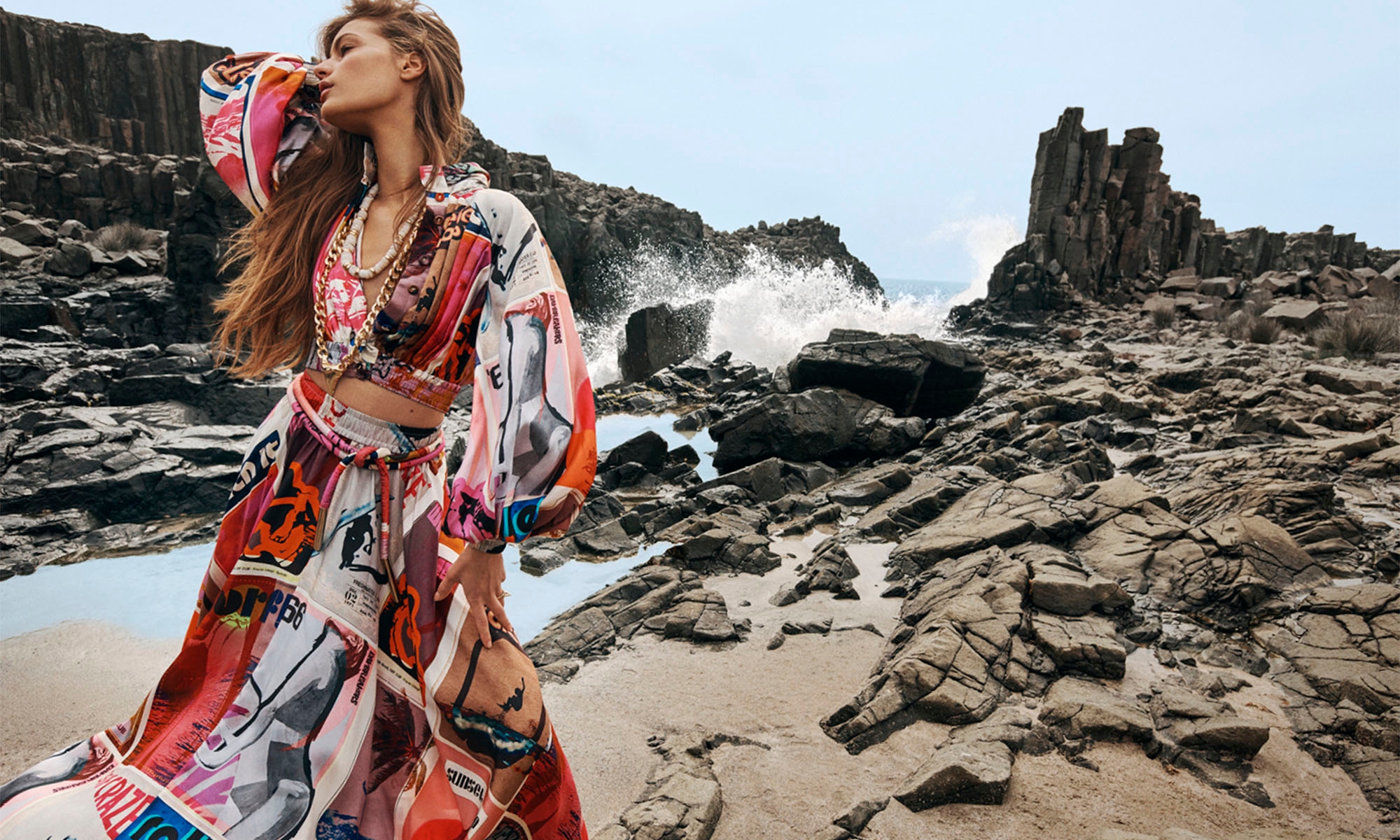 Fashion editorial shot of a young adult female model posing in a colorful patterned dress on a rocky beach near crashing ocean waves.