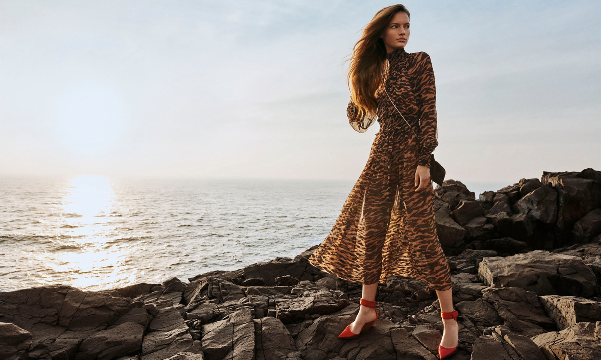 A woman poses on a rocky outcrop in an animal print maxi dress and red heels in a fashion style shot against a sunset and ocean backdrop.