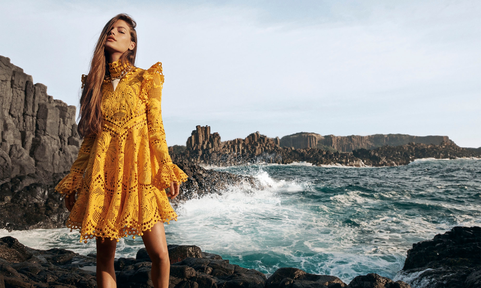 Woman in a yellow lace dress stands on rocks near the ocean with a rocky coastline in the background on a sunny day in an outdoor fashion shot.