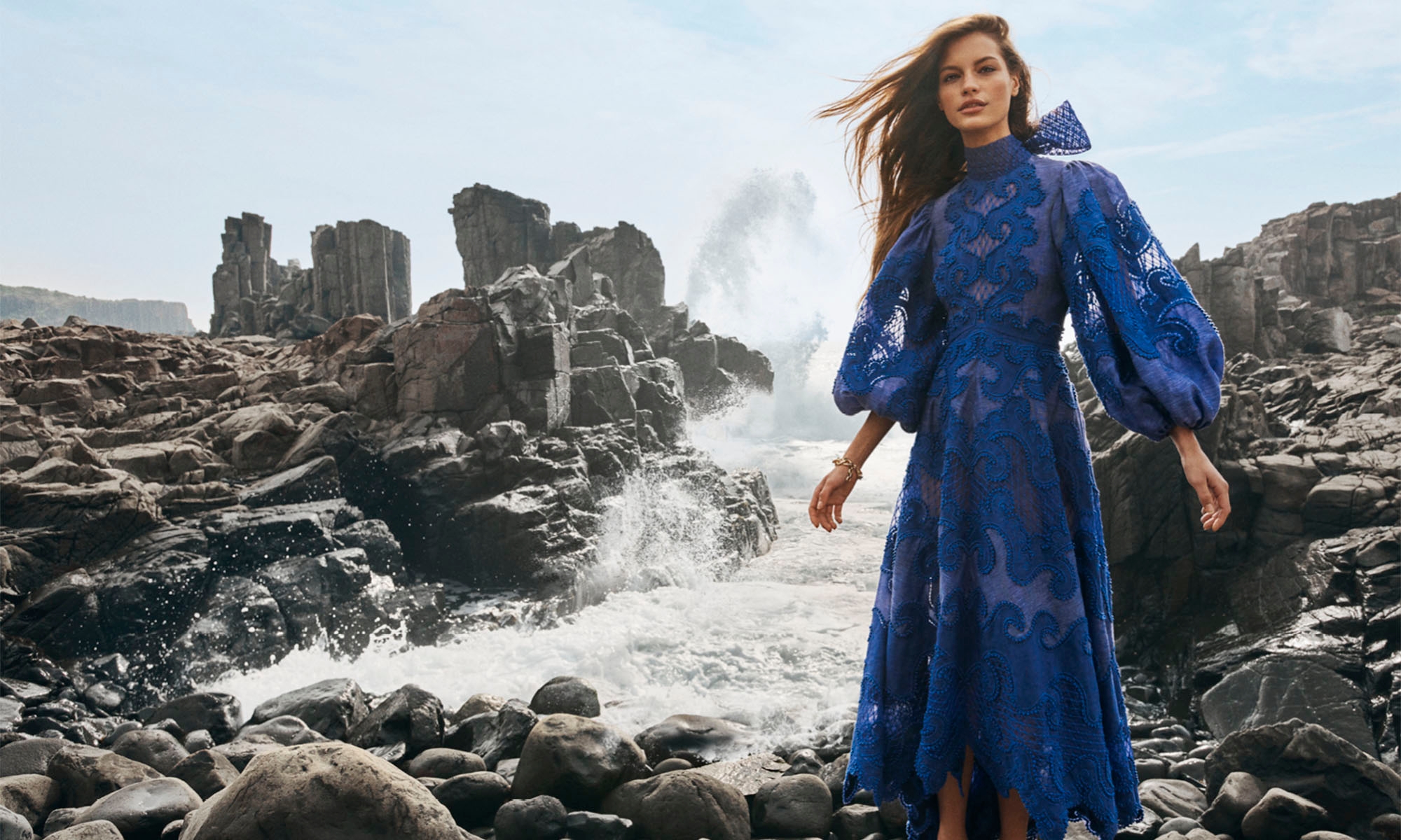 Fashion shot of a woman in a blue lace dress standing on a rocky shore with ocean waves and rock formations in the background.