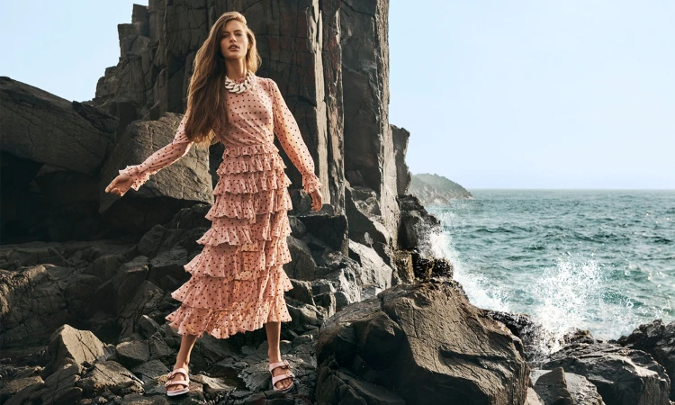 Model in a pink ruffled polka dot dress standing on dark rocky coastal rocks near the crashing ocean for Zimmermann Spring RTW