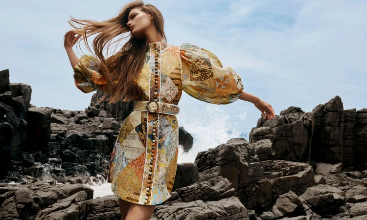 Model with long brown hair wearing a patterned dress with puffed sleeves standing among dark rocky formations