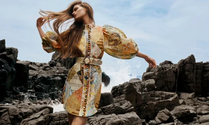 Model with long brown hair wearing a patterned dress with puffed sleeves standing among dark rocky formations