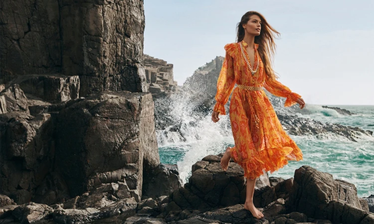 Model wearing a vibrant orange patterned dress posing on rocky coast by the ocean