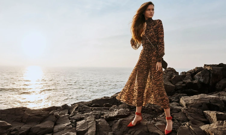 A model wearing a patterned long-sleeve dress and red shoes standing on rocky coastal terrain beside the ocean during sunset.
