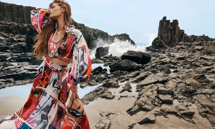 Model wearing a vibrant printed two-piece outfit posing against a rugged rocky coastline with crashing waves.