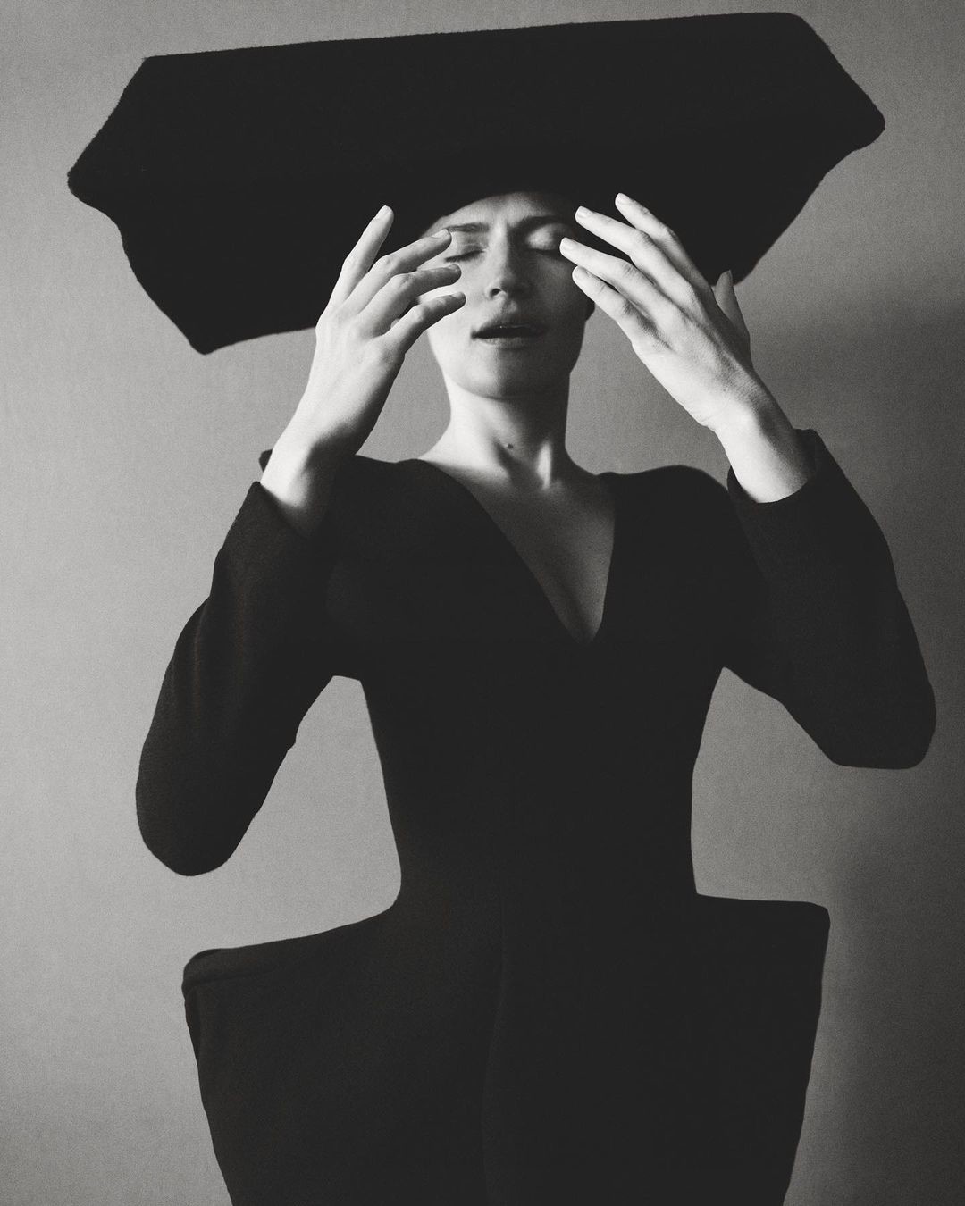 Elegant studio shot of a stylish adult woman with hands near her face, wearing a black dress and a large black hat against a neutral backdrop.