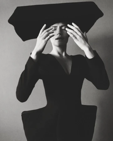 Elegant studio shot of a stylish adult woman with hands near her face, wearing a black dress and a large black hat against a neutral backdrop.