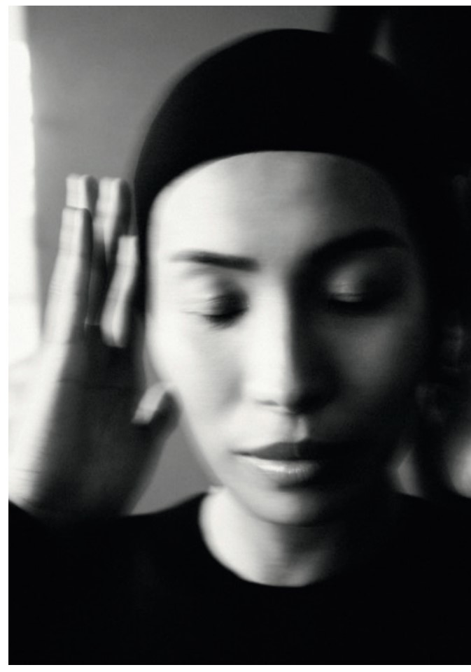 Monochromatic portrait of a woman with dark hair, wearing a black head covering, with her eyes closed and blurred hand near face.
