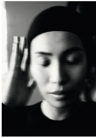 Monochromatic portrait of a woman with dark hair, wearing a black head covering, with her eyes closed and blurred hand near face.