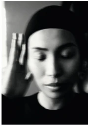 Close-up monochrome portrait of a model with eyes closed wearing a dark cap and raising hand to head.