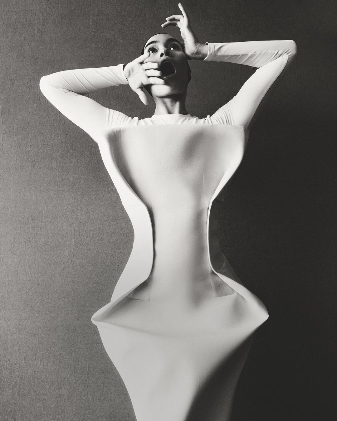 Monochrome fashion shot of a slender woman in a white gown with an open mouth, hands framing her face, set against a plain background.
