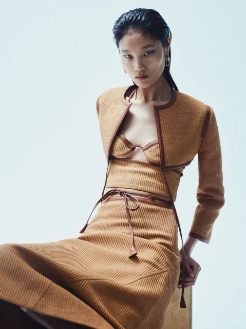 A seated Asian woman poses in a tan ribbed, three-piece skirt suit with brown leather trim against a pale backdrop in a studio shot.