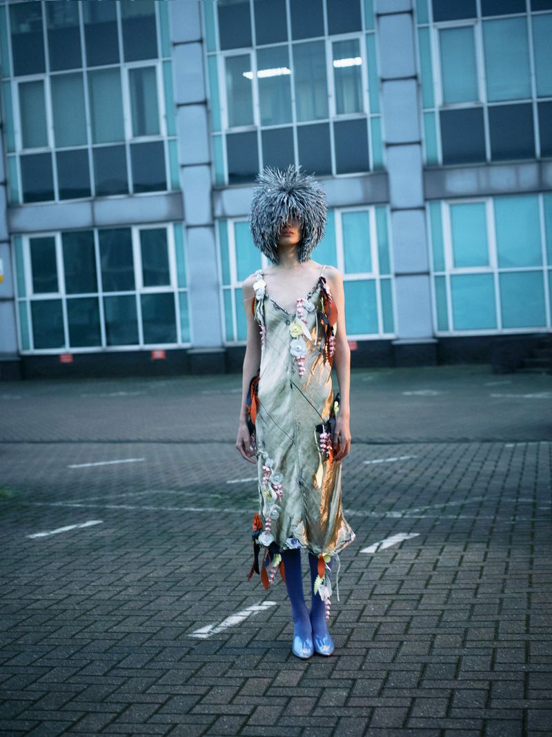Fashionable woman wearing an elaborate, textured gray headpiece and embellished dress, standing on a cobblestone surface in an urban environment.