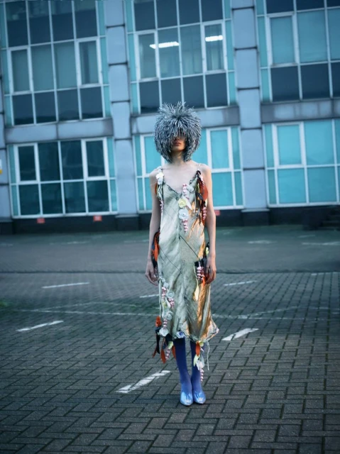 Fashionable woman wearing an elaborate, textured gray headpiece and embellished dress, standing on a cobblestone surface in an urban environment.