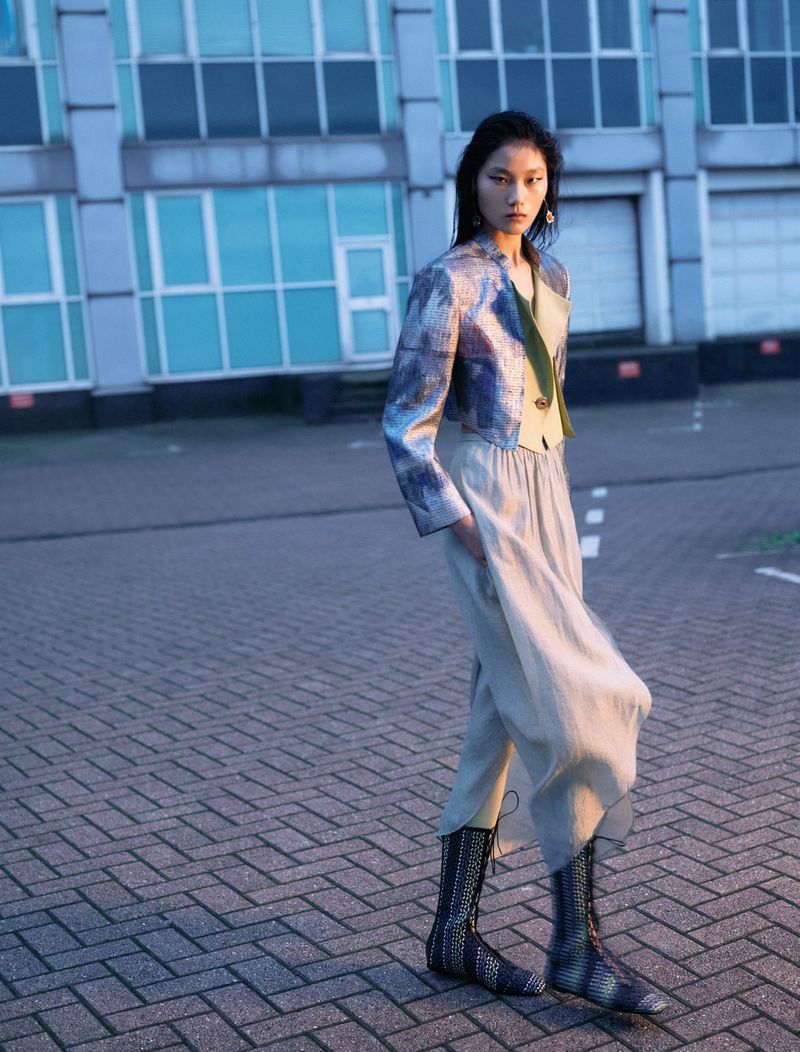 An Asian woman in a blue brocade jacket and wide leg pants stands outside a modern building on a brick-paved area, fashion editorial.