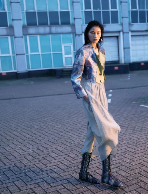 An Asian woman in a blue brocade jacket and wide leg pants stands outside a modern building on a brick-paved area, fashion editorial.