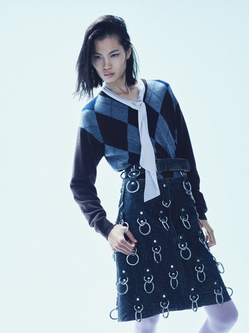 Fashion shot of a woman wearing a patterned sweater, dark denim skirt with metal ring details, and white tights against a white background.