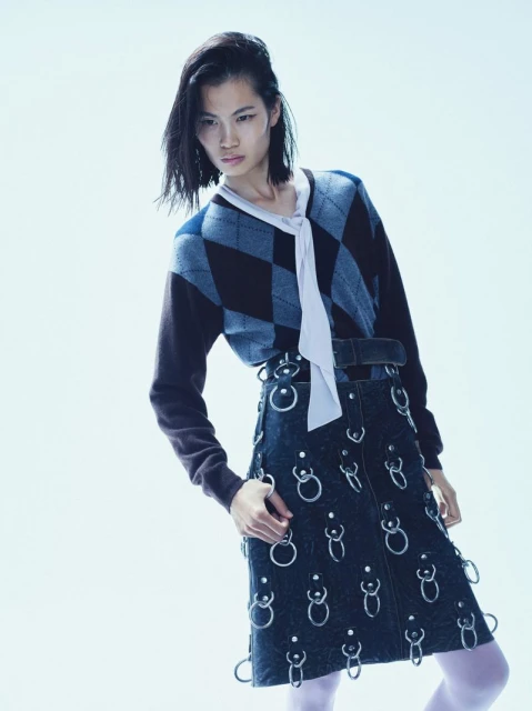 Fashion shot of a woman wearing a patterned sweater, dark denim skirt with metal ring details, and white tights against a white background.