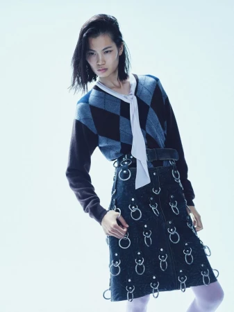 Fashion shot of a woman wearing a patterned sweater, dark denim skirt with metal ring details, and white tights against a white background.