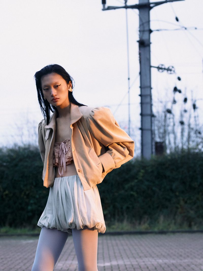Young woman in a tan jacket poses in an outdoor fashion shot, showcasing contemporary style and neutral colors against an urban backdrop.