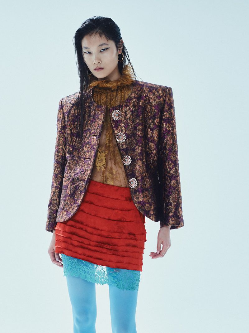 Fashion model poses in a textured purple jacket, red skirt, and blue tights against a white backdrop, showcasing contemporary style.