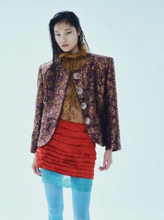 Fashion model poses in a textured purple jacket, red skirt, and blue tights against a white backdrop, showcasing contemporary style.