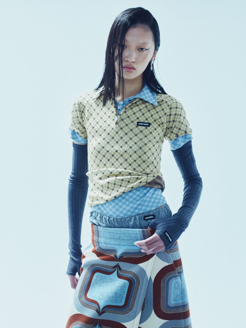 Asian woman posing in layered fashion, wearing a patterned top, blue shirt, gray sleeves, and retro-patterned skirt against a white backdrop.
