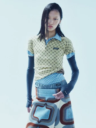 Asian woman posing in layered fashion, wearing a patterned top, blue shirt, gray sleeves, and retro-patterned skirt against a white backdrop.